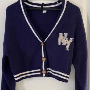 Marinblå croppad kofta från H&M Divided - Snygg marinblå croppad kofta med vita ränder och broderad NY-logga på bröstet. Koftan har v-ringning, långa ärmar och tre träknappar framtill. Stickad i mjukt material och har ribbade detaljer vid krage och nederkant.