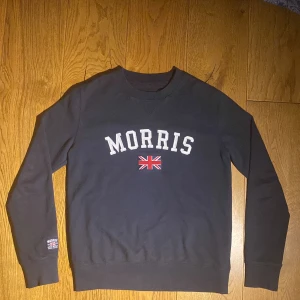 Marinblå sweatshirt från Morris - Säljer en marinblå sweatshirt från Morris med texten 'Morris' och en brittisk flagga på framsidan. Tröjan har långa ärmar och en rund halsringning. Perfekt för en avslappnad stil. Storlek Xs