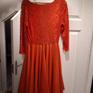 Red spetsklänning  - Snygg orange klänning med v-ringad spetsöverdel och trekvartsärmar. Kjolen är vid och har ett mjukt fall. Spetsen har blommigt mönster och klänningen stängs med dragkedja bak. Perfekt för dig som vill sticka ut med färg och detaljer.