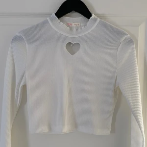 Vit ribbad långärmad topp med hjärtformad detalj - Superfin vit ribbad topp från Romwe i storlek S. Croppad modell med lång ärm och ett hjärtformat utskuret hål vid halsen. Stretchigt material och hög krage ger en snygg och trendig look. Perfekt att styla med jeans eller kjol.