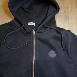 Svart Moncler hoodie med dragkedja - Svart hoodie från Moncler med hel dragkedja framtill, huva med snörning och diskret Moncler-logga på bröstet. Tillverkad i mjuk bomull och har två fickor framtill. Perfekt för en chill och stilren look.