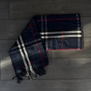 Burberry inspirerad halsduk - Snygg mörkblå halsduk med klassiskt rutigt mönster i vitt, svart och rött. Halsduken har långa fransar i ändarna och känns både stilren och trendig. Perfekt accessoar för att lyfta din outfit under kyliga dagar.
