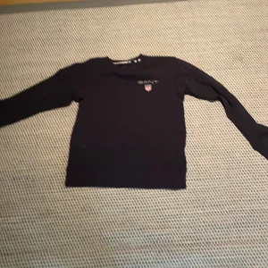 Svart långärmad tröja från GANT - Säljer en svart långärmad tröja från GANT med broderad logga och sköld på bröstet. Tröjan har en klassisk passform och är tillverkad i mjuk bomull, perfekt för en stilren och enkel look.