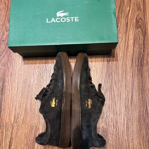 Svarta Lacoste sneakers i mocka - Säljer ett par svarta Lacoste Baseshot sneakers i mocka med gummisula och guldfärgad krokodillogga på sidan. Klassisk låg modell med snörning och diskret branding på plösen. Perfekta för dig som gillar stilrena och sportiga skor. Använda max 3 månader