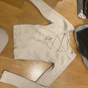 Ljus ribbad långärmad topp med snörning - Säljer en ljus, ribbad långärmad topp med snygg snörning framtill i v-ringad design. Toppen har en croppad passform och är tillverkad i mjukt material som sitter skönt. Perfekt för dig som gillar trendiga detaljer och stilren look.