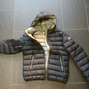 Svart dunjacka med huva från Colmar - Snygg svart dunjacka från Colmar med huva och två dragkedjeförsedda fickor. Jackan har quiltad design och Colmar-logga på ärmen. Insidan är ljusgrå och jackan stängs med en robust dragkedja. Perfekt för kyliga dagar och stilren look. Mindre skador (se bilder) påverkar deremot inte jackan på något sätt och syns inte derför lite lägre pris. Annars i bra skick perfekt nu till vintern/hösten. 