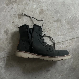 Svarta boots från Wild Falls - Svarta boots från Wild Falls med snörning och dragkedja på sidan. Skorna har en ljus, grov sula och detaljer i brunt skinn baktill. Tillverkade i syntetmaterial med rund tå och platt sula. Perfekta för dig som gillar en robust och stilren look.