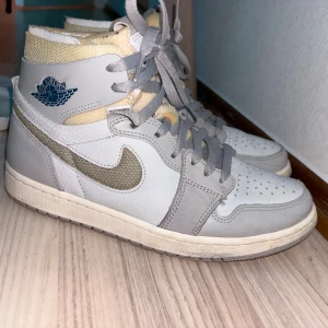 Nike Air Jordan 1 High grå/vit - Säljer ett par Nike Air Jordan 1 High i grått och vitt med beige detaljer. Skorna har klassisk snörning, rund tå och är tillverkade i skinn och textil. Swoosh-loggan är i mesh och Air Jordan-loggan syns på sidan. Perfekta för dig som gillar streetwear och sneakers med ikonisk design.