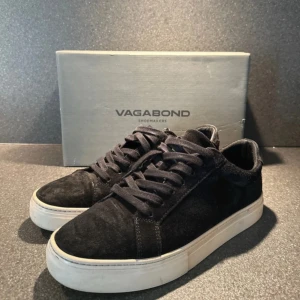 Svarta mockasneakers från Vagabond - Snygga svarta sneakers från Vagabond i mjuk mocka med klassisk vit platt sula. Skorna har rund tå och snörning framtill för en clean och stilren look. Perfekta för dig som gillar minimalistisk stil och vill ha ett par bekväma vardagssneakers.
