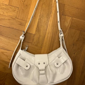 Vit axelväska med krokodilmönster - Snygg vit axelväska med krokodilmönstrad yta och justerbar rem. Väskan har silverfärgade detaljer, två fickor med knappar framtill och en dragkedja upptill. Perfekt storlek för mobilen och det viktigaste. Ger en clean och trendig vibe till din outfit.