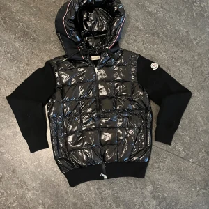 Svart Moncler pufferjacka med huva - Snygg svart pufferjacka från Moncler med glansig quiltad front och huva. Ärmarna är stickade och jackan har dragkedja framtill. Ikonisk Moncler-logga på ärmen och sportig känsla. Perfekt för kyliga dagar när du vill ha både stil och värme. PRIS KAN DISKUTERAS VID SNABB AFFÄR
