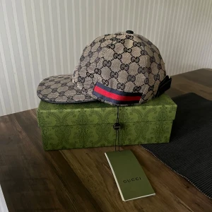 Gucci keps med GG-mönster och röd/blå detalj - Snygg keps från Gucci med klassiskt GG-mönster i beige och svart. Kepsen har en böjd skärm och en ikonisk röd/blå rand på sidan. Tillverkad i bomull med justerbar rem bak. Perfekt för dig som vill ha en lyxig och trendig accessoar.
