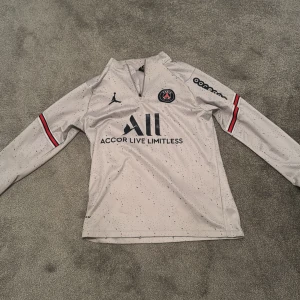 PSG långärmad fotbollströja M - Säljer en PSG långärmad fotbollströja i ljusgrått med svarta prickar, röda och svarta detaljer på ärmarna. Tröjan har Jordan-logga, PSG-emblem och tryck som 'Accor Live Limitless' och 'Visit Rwanda'. Tillverkad i mjukt, tekniskt material som andas.