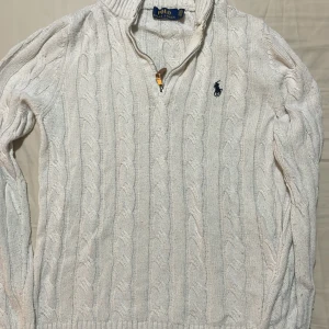 Vit kabelstickad tröja Polo Ralph Lauren - Vit kabelstickad tröja från Polo Ralph Lauren med halv dragkedja och broderad marinblå logga på bröstet. Tröjan har hög krage, långa ärmar och ribbade muddar. Perfekt för lager-på-lager under höst och vinter. OBS! Pris kan diskuteras. 