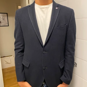 Mörkblå kavaj med två knappar - Snygg mörkblå kavaj med två knappar framtill och klassiska fickor med lock. Kavajen har smala slag och en bröstficka, perfekt för en stilren look. Passar till skjorta eller t-shirt för en modern vibe.