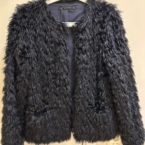  Mörk blå pälsjacka från Zara Basic - Säljer en  pälsjacka från Zara Basic med fluffig och långhårig struktur. Jackan har rund halsringning, två fickor framtill och är helsvart. Perfekt för dig som vill sticka ut med en cool och trendig look.