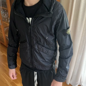 Svart vindjacka från Stone Island - Säljer en svart vindjacka från Stone Island med huva och flera snedställda fickor med dragkedja. Jackan har den klassiska logotypen på ärmen och är tillverkad i vattenavvisande och andningsbar nylon. Perfekt för dig som gillar streetwear och snygga detaljer. Modellen är 186, väger 70 som ni ser är den rätt tajt så denna passar dig som är lite mindre o kortare. 