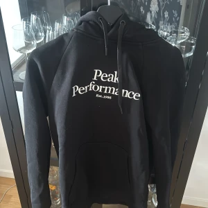 Svart hoodie Peak Performance - Svart hoodie från Peak Performance med vit logga på bröstet. Klassisk modell med huva och dragsko, samt stor magficka framtill. Perfekt för chill dagar eller när du vill ha en clean streetstyle-look.
