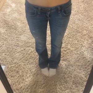 Blå bootcut jeans med slitningar - Säljer ett par blå bootcut jeans med snygga slitningar på låren och klassiska fem fickor. Jeansen har låg midja och är tillverkade i mjukt denimtyg som sitter skönt. Perfekta för dig som gillar en avslappnad och trendig look.