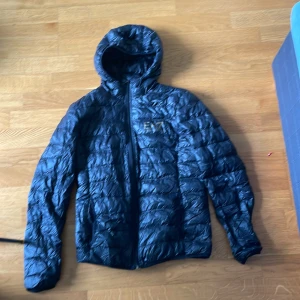 Svart pufferjacka EA7 Emporio Armani - Snygg svart pufferjacka från EA7 Emporio Armani med guldigt logotryck på bröstet och matchande dragkedja. Jackan har huva, quiltad design och lång ärm. Perfekt för dig som gillar stilrena och moderna jackor med exklusiv känsla. Pris kan diskuteras 