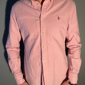 Rosa skjorta från Ralph Lauren - Klassisk rosa skjorta från Ralph Lauren med button-down krage och broderad logga på bröstet. Modellen är 177-178cm, Skjortan har lång ärm, normal passform och är tillverkad i mjuk bomull. Perfekt för en clean och stilren look. 