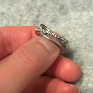 Silverfärgad ring med glittrande stenar - Snygg ring i silverfärg med ett coolt korsat band och glittrande stenar längs ena sidan. Ringen har en modern och unik design som sticker ut och passar perfekt för dig som gillar att glänsa lite extra.