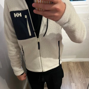 Vit fleecejacka från Helly Hansen - Säljer en vit fleecejacka från Helly Hansen med marinblå bröstficka och dragkedjor. Jackan har huva, hel dragkedja framtill och två sidofickor med dragkedja. Perfekt för kyliga dagar och riktigt skön att ha på sig. Inga fläckar eller märken.