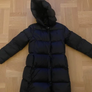 Svart lång dunjacka Polo Ralph Lauren - Lång svart dunjacka från Polo Ralph Lauren med huva och dragkedja framtill. Jackan är quiltad med breda kanaler och har en diskret logga broderad på bröstet. Perfekt för kalla dagar när du vill hålla värmen och ändå se stilren ut. BARN STORLEK PASSAR 10 ÅR