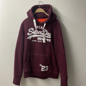 Vinröd hoodie från Superdry - Säljer en vinröd hoodie från Superdry med vit tryckt logga och japanska tecken på bröstet. Tröjan har huva med vita snören, känguruficka och ribbade muddar. Perfekt för dig som gillar streetwear och vill ha en chill look. Barn storlek L men motsvarar herr S.