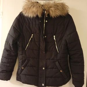 Svart pufferjacka med fuskpäls från H&M - Svart pufferjacka från H&M i storlek 40 med fluffig fuskpäls på huvan. Jackan har quiltad design, två snedställda fickor med dragkedja och tryckknappar framtill. Materialet är polyester och akryl, perfekt för kalla dagar. Snygg och varm look för vintern.