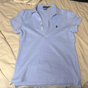 Ljusblå pikétopp från Polo Ralph Lauren - Säljer en ljusblå pikétopp från Polo Ralph Lauren med klassisk krage och korta ärmar. Toppen har knappar framtill och den ikoniska broderade loggan på bröstet. Tillverkad i mjuk bomull som är skön mot huden. Perfekt för en chill och stilren look. 
