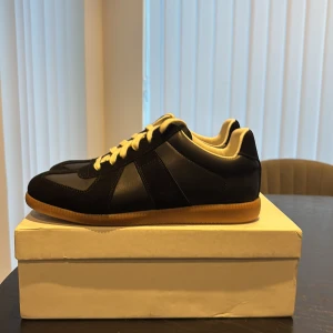 Maison Margiela svarta gats  - Maison Margiela sneakers i svart skinn och mocka med vita snören och beige gummisula. Klassisk rund tå och platt sula, stilren design med kontrasterande material och färger. Perfekt för dig som gillar minimalistisk och trendig stil.