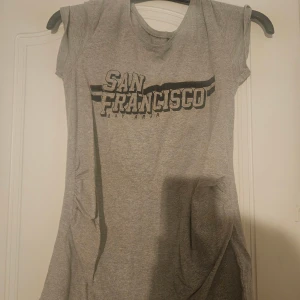 Grå t-shirtklänning San Francisco - Grå t-shirtklänning med tryck 'San Francisco Bay Area' framtill. Klänningen har korta ärmar och är tillverkad i mjuk bomull med lite stretch för extra komfort. Enkel och avslappnad stil, perfekt för en chill look. Mammatopp