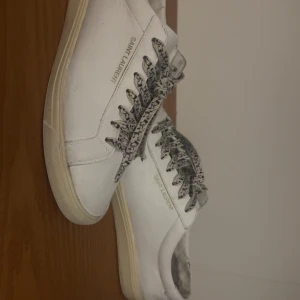 Vita Saint Laurent sneakers i skinn - Säljer ett par klassiska vita sneakers från Saint Laurent i mjukt skinn. Skorna har rund tå, platt sula och coola snören med svart mönster. Diskret Saint Laurent-logga på sidan och bak på sulan. Perfekta för en clean och stilren look.