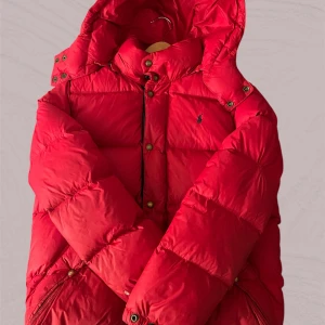 Röd dunjacka från Polo Ralph Lauren - Säljer en röd dunjacka från Polo Ralph Lauren med huva och dragkedja samt knappar framtill. Jackan har två snedställda fickor med tryckknappar och klassisk Polo-logga broderad på bröstet. Perfekt för alla dagar. Den är i 7/10 skick.
