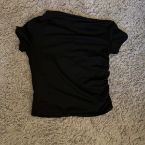 Svart off shoulder t shirt  - En svart, kortärmad topp med snygg rynkdetalj längs ena sidan. Toppen har en enkel och stilren design med rund hals och är tillverkad i ett mjukt, stretchigt material som sitter skönt på kroppen.