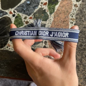 Blå Christian Dior J'Adior armband - Snyggt blått armband från Christian Dior med texten 'J'Adior' broderad i vitt. Armbandet har en vävd design med vita detaljer och avslutas med två coola tofsar. Perfekt accessoar för att lyfta din outfit med lite lyxig vibe.