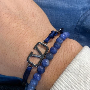 Blått Valentino armband med pärlor - Snyggt armband från Valentino med blå snöre och en metallfärgad V-logga. Kombinerat med ett pärlarmband i blå toner som ger en cool och modern vibe. Perfekt för dig som gillar stilrena accessoarer med designer-touch.