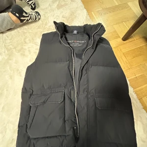 Svart dunväst från Gant Sport - Svart dunväst från Gant Sport med hög krage och två stora fickor framtill. Västen har quiltad design och stängs med dragkedja. Perfekt för lager-på-lager och ger en clean, sportig look.