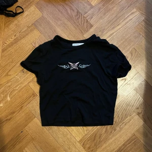 Svart croppad topp från Bershka - Svart croppad topp från Bershka med korta ärmar och ett snyggt tribal-inspirerat broderi i ljusrosa och ljusblått framtill. Mjuk bomullskvalitet och rund halsringning. Perfekt för dig som gillar edgy och trendiga detaljer.
