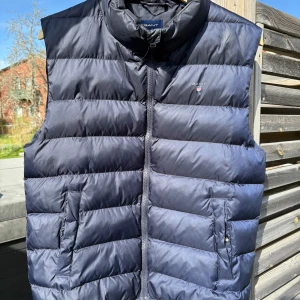 Mörkblå dunväst från Gant - Snygg mörkblå dunväst från Gant med dragkedja framtill och två fickor med knappar. Västen har en klassisk quiltad design och hög krage. Perfekt för våren och lite kyligare sommarkvällar                              Nypris: ca 1500kr                                                    pris kan diskuteras vid snabb affär 