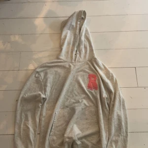 Grå hoodie med Rosa  patch - Snygg grå hoodie med huva och en rosa patch med drip-effekt på bröstet. Tröjan har en klassisk känguruficka framtill och långa ärmar. Perfekt för chill dagar och streetwear-stil. Knappt använd 