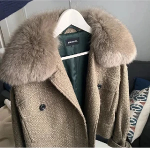 Beige pälsjacka från Meotine S/M - Säljer en beige pälsjacka från Meotine med stor fluffig krage i äkta rävpäls. Jackan har två stora fickor framtill, knappar och en croppad, loose passform. Materialet är en mix av ull och polyester med fodrad insida. Perfekt statement-plagg för höst och vinter.