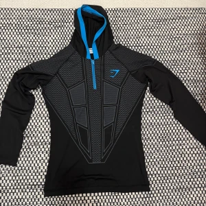 Gym Shark V1  - Sportig svart hoodie från Gymshark med coolt geometriskt mönster i grått och blått. Halv dragkedja och blå insida på huvan ger en snygg kontrast. Perfekt för träning eller chill, med långärmad passform och tekniskt syntetmaterial.