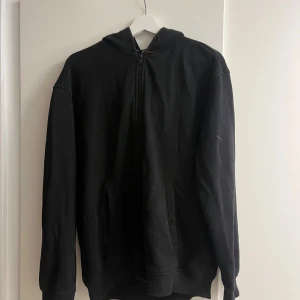 Svart hoodie med dragkedja från H&M - Svart hoodie från H&M i relaxed fit med huva och hel dragkedja framtill. Tröjan har långa ärmar, två fickor och är gjord i mjuk bomullsmix. Perfekt för chill dagar och enkel att matcha med jeans eller joggers.