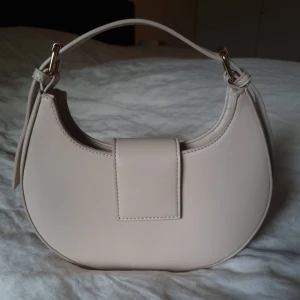 Public Desire axelväska  - The Irie Stone PU Circular Buckle Strap Grab Bag från Public Desire i neutral färg. Axelrem kan justeras. Spåret av användning finns. Mått 23 x 18 x 7 cm