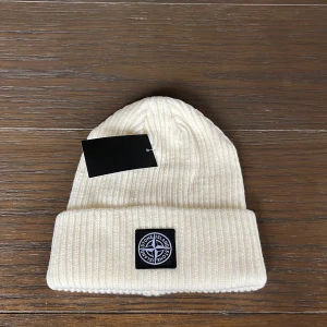 Vit ribbad mössa från Stone Island - Snygg vit ribbstickad mössa från Stone Island med klassisk logopatch framtill. Mössan har uppvikt kant och är tillverkad i mjukt stickat material som håller dig varm och stilren under kyliga dagar.