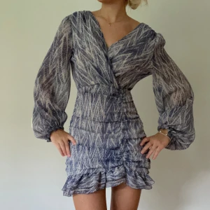 Blå mönstrad omlottklänning med volanger - Supersnygg kort omlottklänning i blått och vitt med unikt randigt mönster. Klänningen har långa puffärmar, v-ringad hals och volangdetaljer nedtill. Materialet är lätt och luftigt, perfekt för varmare dagar. Sitter figurnära över midjan och ger en riktigt fräsch siluett. (Knapp trasig ibak!)