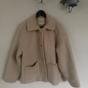 Beige teddyjacka från Monki - Supermysig beige teddyjacka från Monki med stora fickor framtill och klassisk krage. Jackan har en loose passform och stängs med knappar. Perfekt för att hålla sig varm och snygg under kyliga dagar.