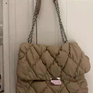 Beige quiltad axelväska med kedja - Säljer en beige quiltad axelväska med silverfärgad kedja som axelrem. Väskan har ett snyggt rutmönster, lock med metallspänne och flera fack inuti. Perfekt storlek för att få plats med allt du behöver och ger en  classy vibe till din outfit.